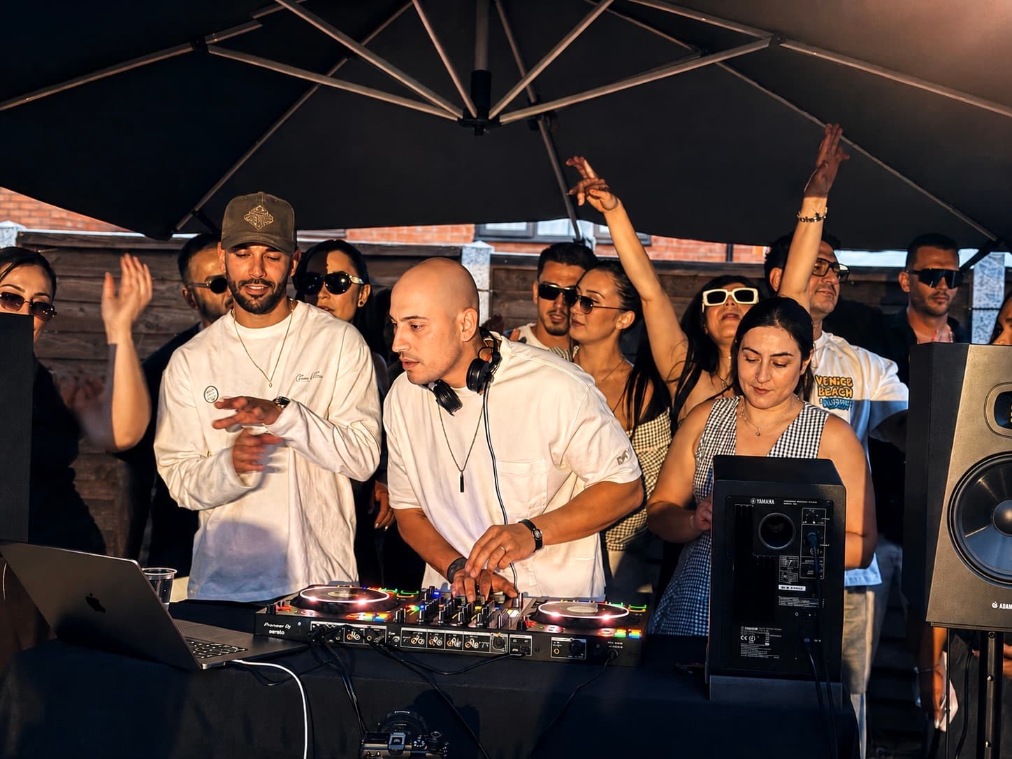 Lostest mixing on a controller at an outdoor party, headphones round his neck, the crowd close behind with hands in the air
